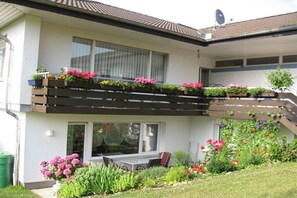 Ein zweigeschossiges Haus mit Balkon, einer Terrasse mit Tisch und Stühlen sowie einem Garten mit verschiedenen Pflanzen und Blumen.