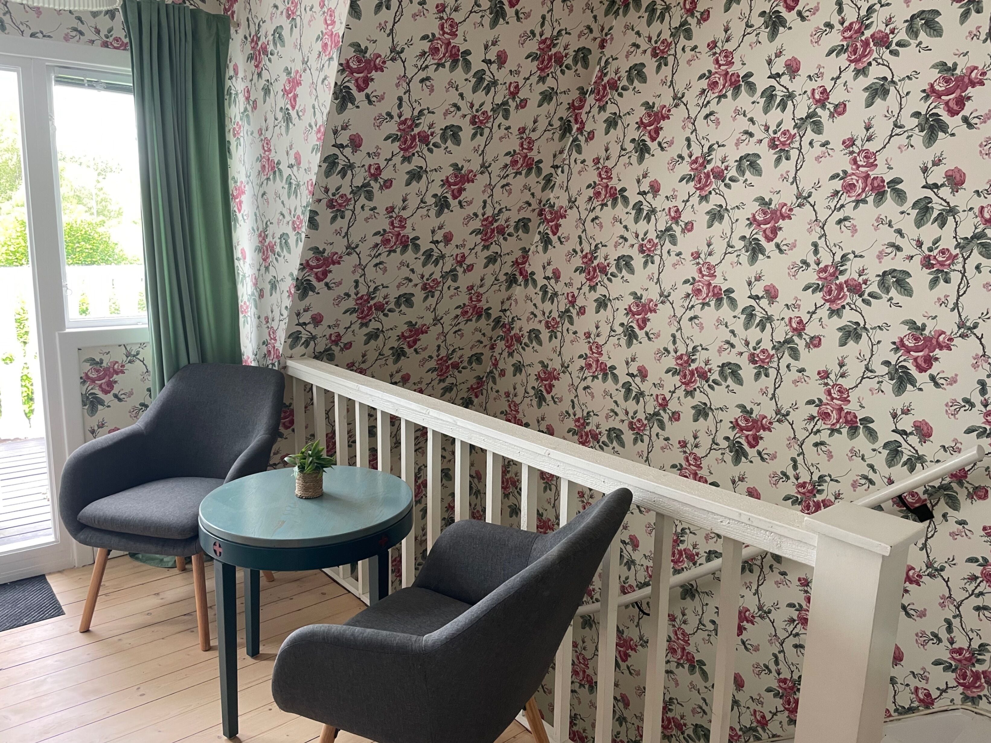 A room with a wooden floor, two grey armchairs, a small round table, a white railing, and floral wallpaper.