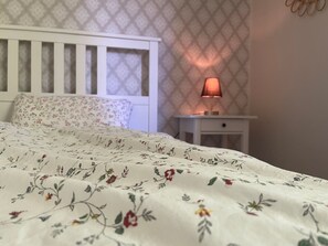 Une chambre à coucher avec une couvre-lit à motifs floraux, une tête de lit blanche, une table de chevet avec une lampe et un papier peint à motifs.