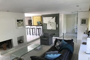 Ein modernes Wohnzimmer mit einem Glastisch, einem Ledersofa mit Kissen, einem grauen Sessel und einem großen abstrakten Gemälde an der Wand.