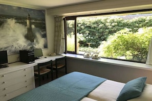 Ein Schlafzimmer mit einem Bett, einem Schreibtisch mit Computer, einem Fenster mit Blick auf Grünflächen und einem Gemälde eines Leuchtturms.