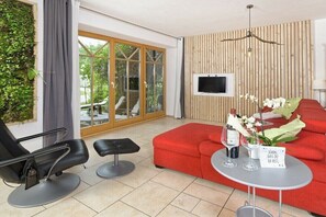 Una sala de estar moderna con un sofá rojo, una silla negra, una mesita con un jarrón y copas de vino, y un televisor montado en una pared de madera.
