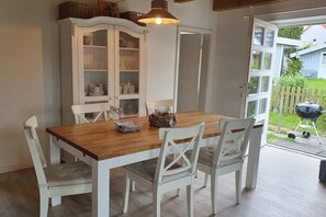 Ein Essbereich mit einem Holztisch, weißen Stühlen, einer weißen Kommode mit Glastüren und Blick in den Garten durch eine offene Tür.