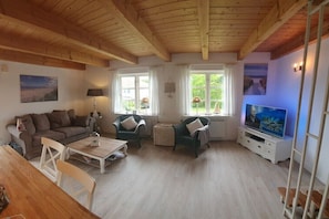 Ein Wohnzimmer mit Holzbalkendecke, einer Couch, zwei Sesseln, einem Couchtisch, einem Fernseher und Fenstern mit Vorhängen.
