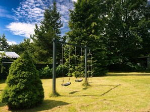 Ein Spielplatz mit Schaukeln in einem grasbewachsenen Garten, Bäume und ein Gebäude im Hintergrund.