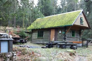 Eine Blockhütte mit einem moosbewachsenen Dach, einer Holzterrasse und einem Steinschild im Vordergrund.