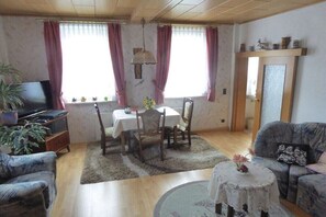 Un salon comprenant un téléviseur, une table à manger, des chaises, un canapé, une petite table ronde ornée de fleurs et une plante en pot.