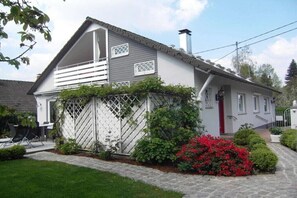 Ein zweigeschossiges Haus mit weißer Fassade, einer roten Tür und einem kleinen Garten mit roten Blumen.