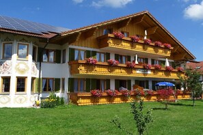 Ein zweistöckiges Hotel mit einem mit Blumen geschmückten Balkon, einem Schild mit der Aufschrift 'Hotel Alpenblick' und einer Solaranlage auf dem Dach.