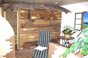 Eine rustikale Holzwand, ein gestreifter Stuhl, ein kleiner Tisch mit einer Blumenkorb und Blick auf eine Landschaft durch ein Fenster.