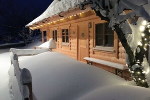 A log cabin with a snow-covered roof, windows, and a bench outside.