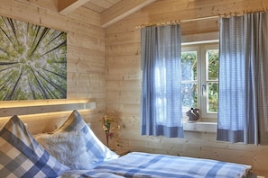 A wooden cabin room with a bed, checkered curtains, a window with a view of greenery, and a wall-mounted painting.