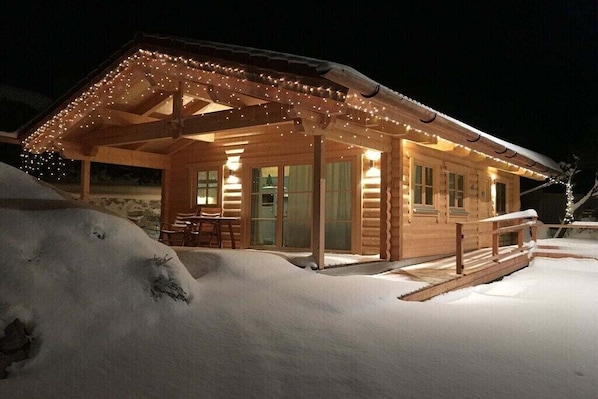 A wooden cabin with a covered porch, a dining area, and a snow-covered landscape at night.