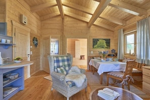 A wooden cabin interior with a dining area, a wicker chair, a table with a white tablecloth, and a clock on the wall.