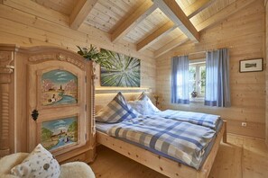 A wooden cabin bedroom with a bed, two paintings, a window with curtains, and a door with a carved design.