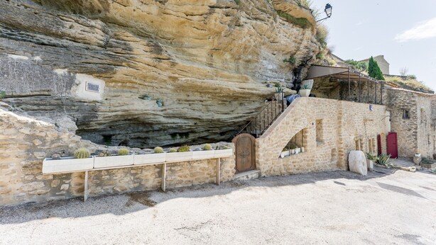 Un edificio de piedra con una escalera que lleva a un nivel superior, un banco rodeado de cactus y una puerta roja.