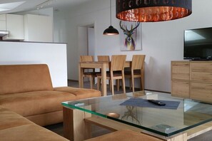 Ein modernes Wohnzimmer mit Glastisch, einer Couch, einer Essecke mit Holzkohlen und einem Fernseher, sowie einem Wanddekor.