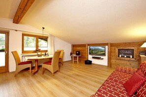 Ein gemütliches Zimmer mit Holzbalkendecke, einem Essbereich mit Korbstühlen, einem Bett mit gemusterten Betttextilien und einem am Wand montierten Fernseher.