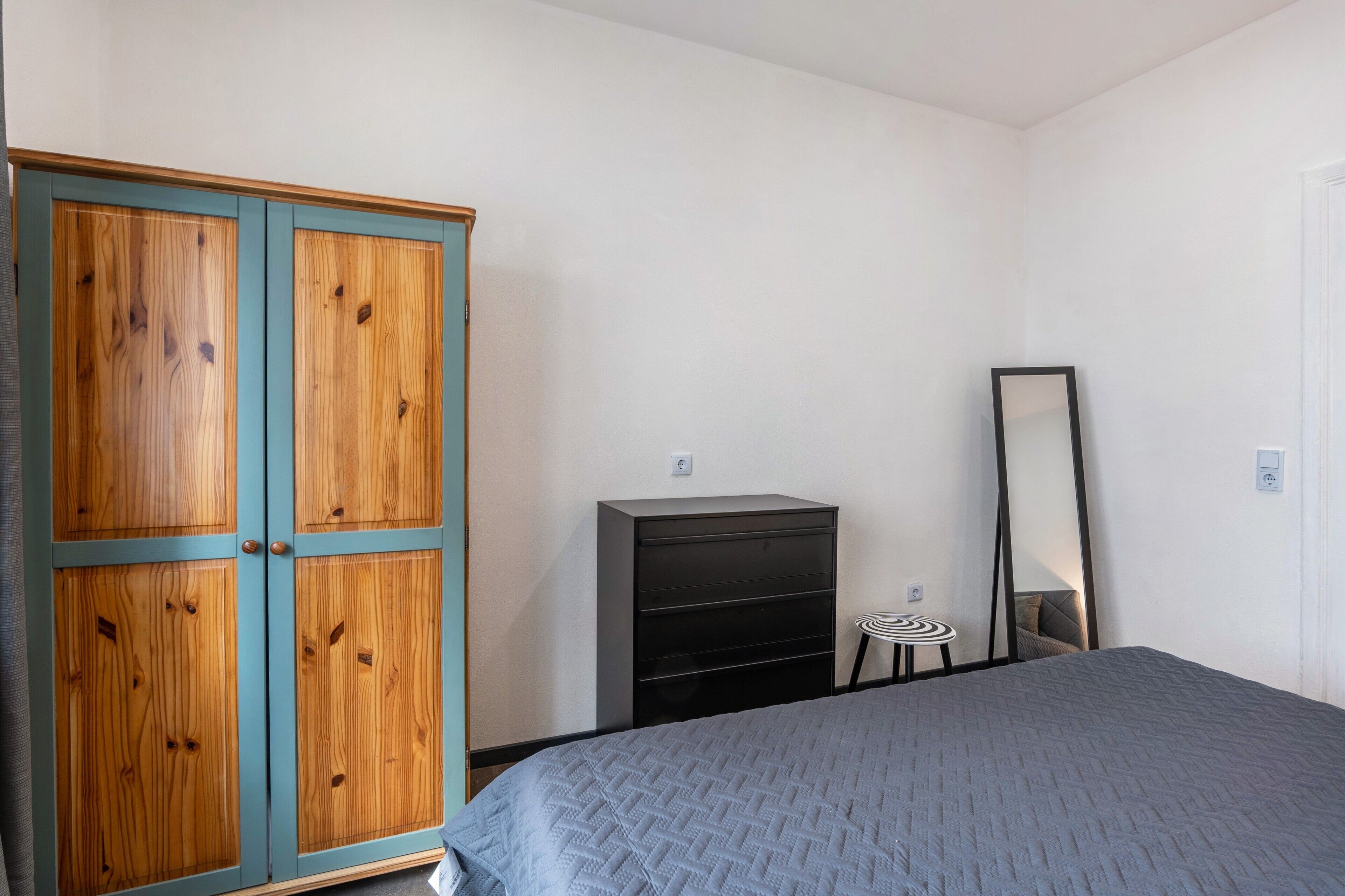 Un dormitorio con cama, armario, cómoda, espejo y un taburete pequeño.