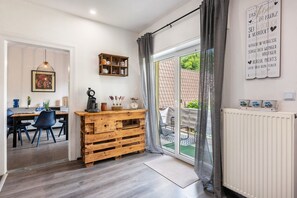 Een kamer met een houten bureau, stoelen, een hanglamp, een plank met spullen en een raam met gordijnen.