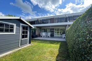 A modern two-story house with a covered patio, a small garden, and a large hedge. - Holiday apartment with 1 bedroom (Petten)