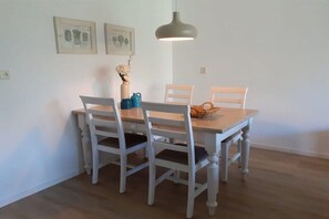 A dining area with a wooden table and four white chairs, a hanging pendant light, and two framed botanical prints on the wall. - Holiday apartment with 1 bedroom (Petten)