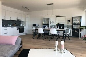 Eine moderne Küche mit weißen Schränken, ein Essbereich mit weißem Tisch und Stühlen sowie eine Wohnbereich mit grauem Sofa.