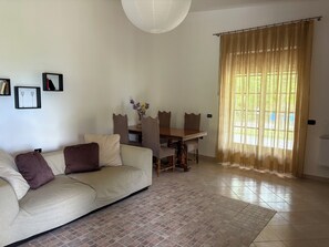 Ein Wohnzimmer mit einer Couch, einem Esstisch mit Stühlen, einer Chandelier und einem Fenster mit Vorhängen.