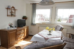 A room with a wooden cabinet, a television, a dining table with a floral tablecloth, and a window with curtains. - Holiday apartment in Esens with swimming pool (Esens)