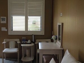 A room with a window, a table, two chairs, a small shelf with bottles, and a mirror on the wall.