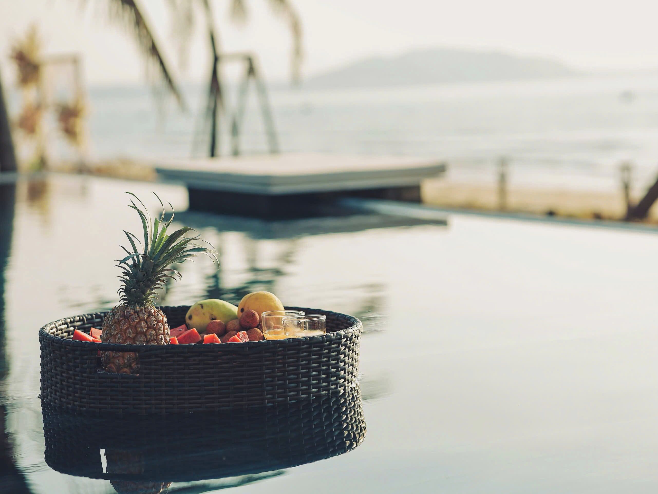 Una cesta de mimbre con frutas y una piña sobre una mesa junto a la piscina.