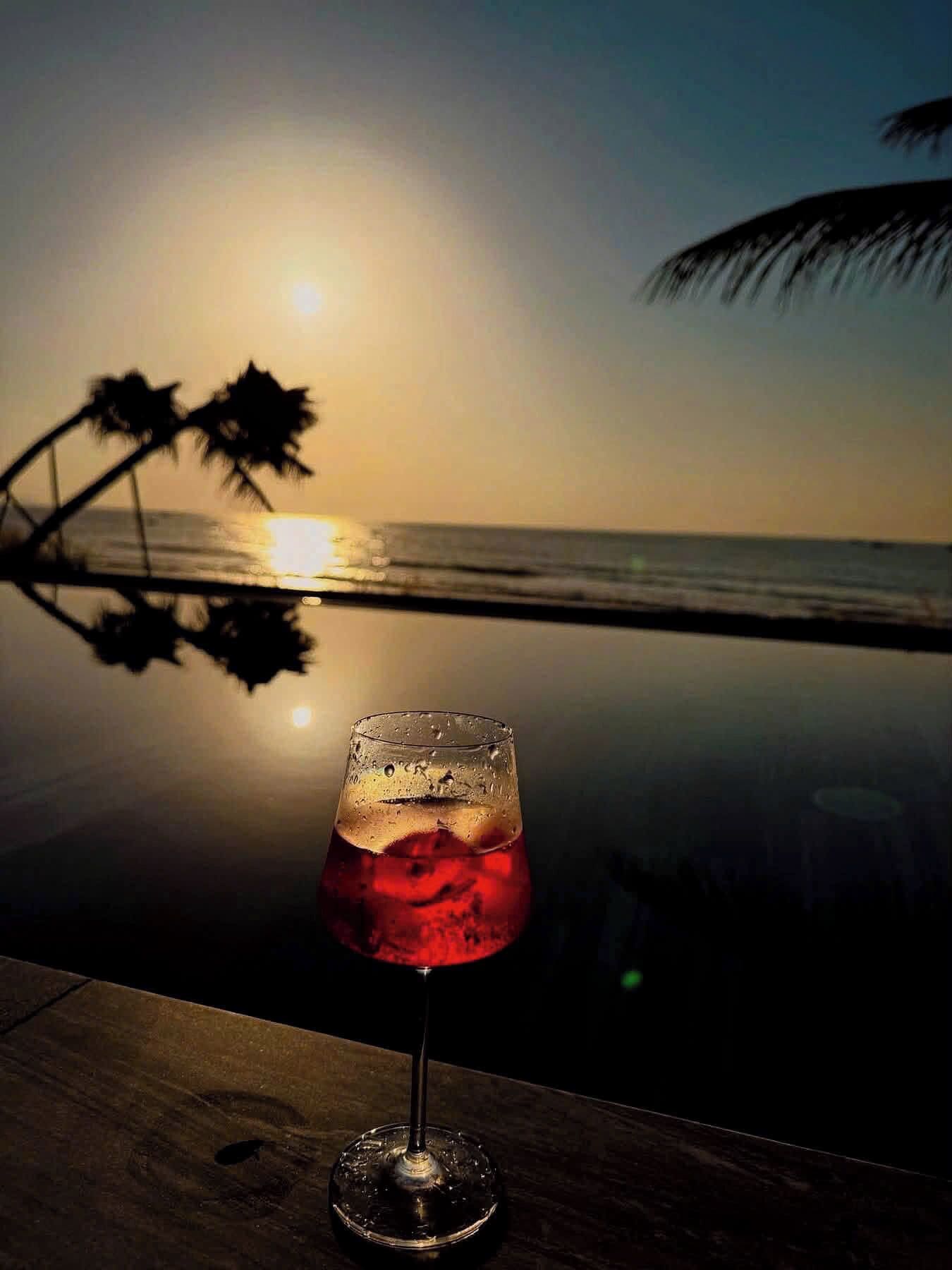 Un vaso de bebida roja sobre una superficie reflectante, con un atardecer y palmeras al fondo.