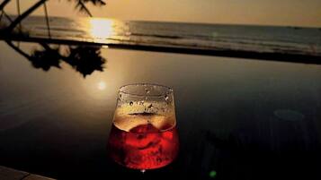 Un vaso de bebida roja sobre una superficie reflectante, con un atardecer y palmeras al fondo.