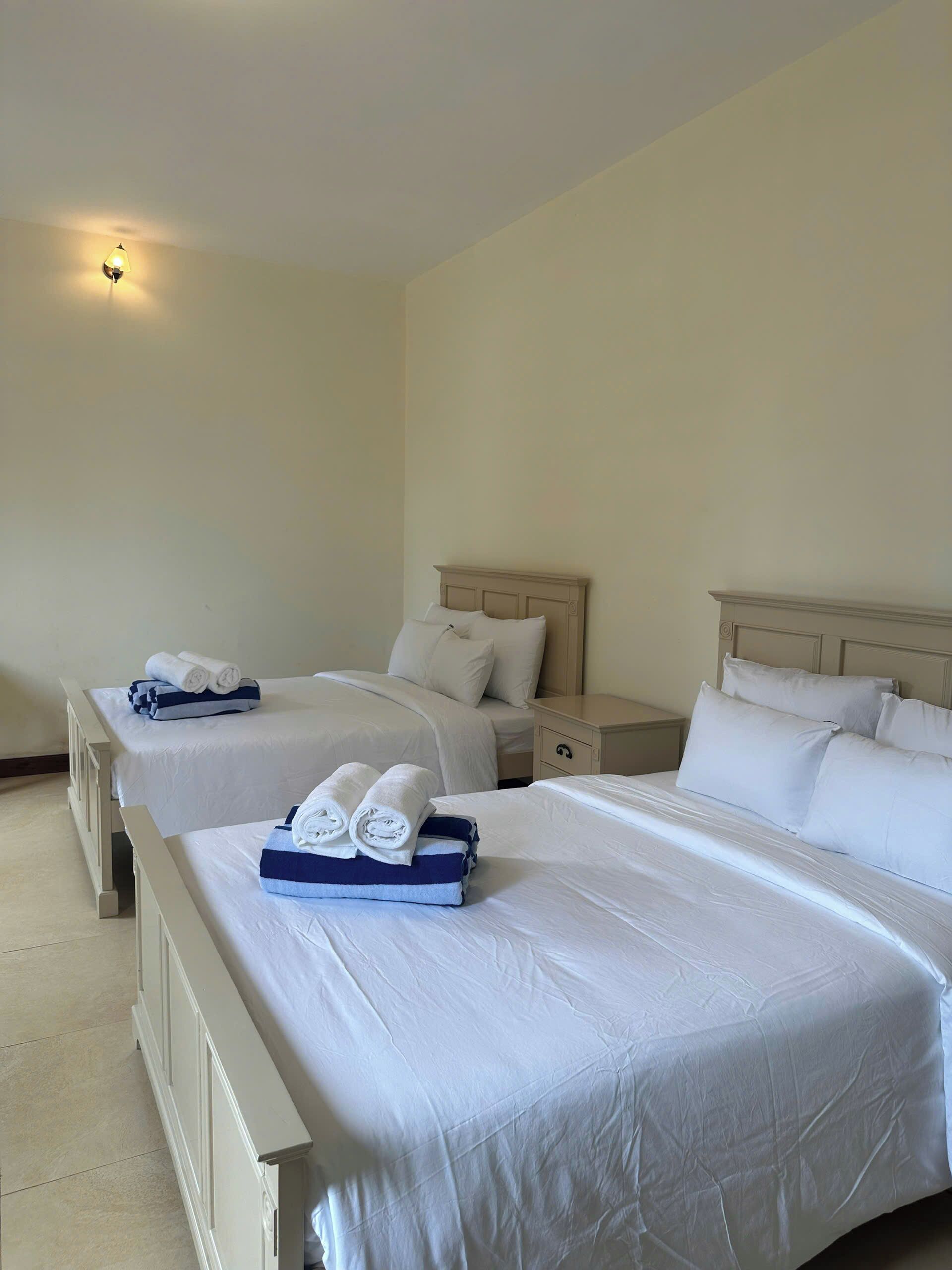 A hotel room with two beds, each with white linens and towels, a nightstand, and a wall-mounted light.