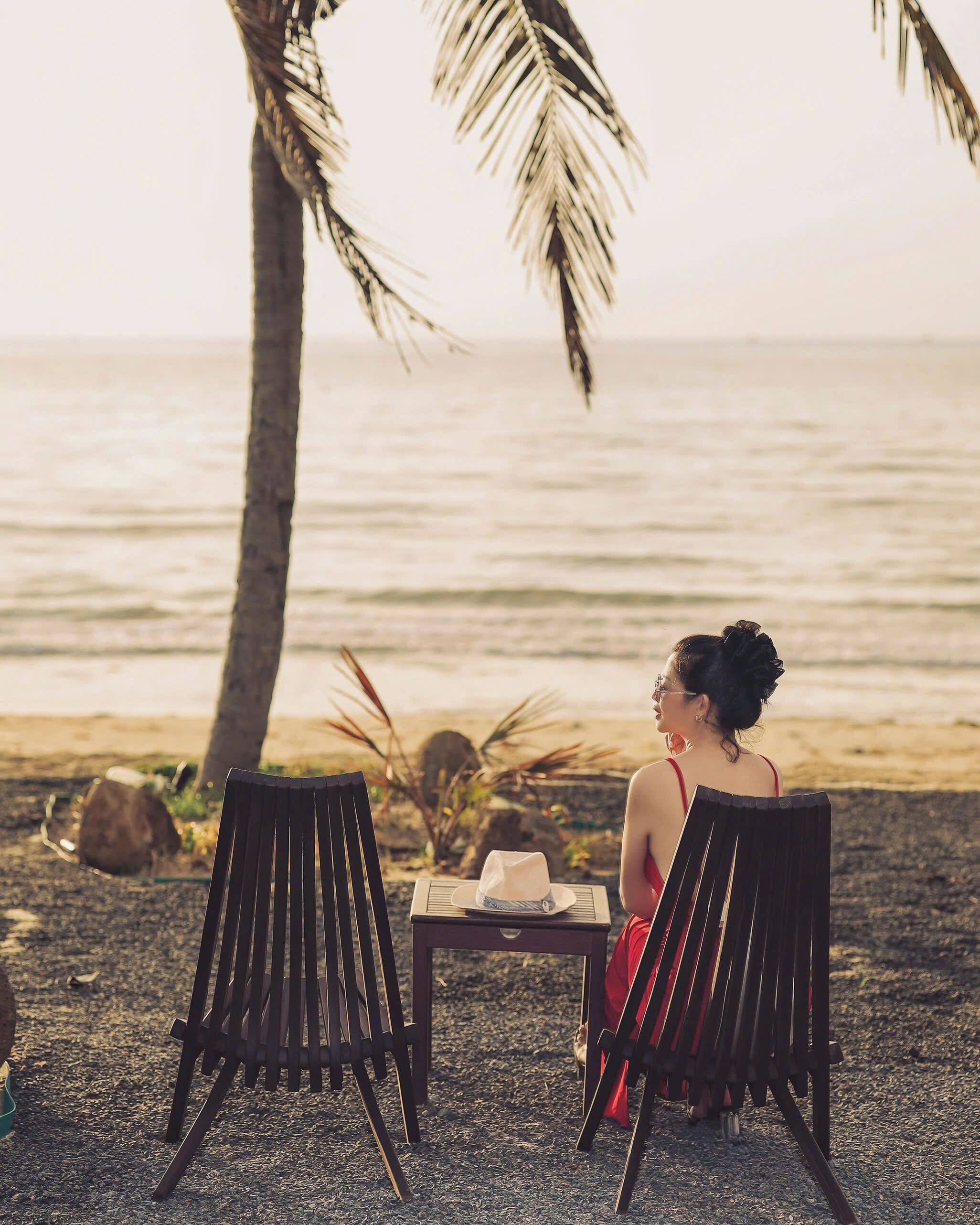 Dos sillas frente a una mesa con un sombrero y una botella, con vista a una playa con palmeras y el océano.