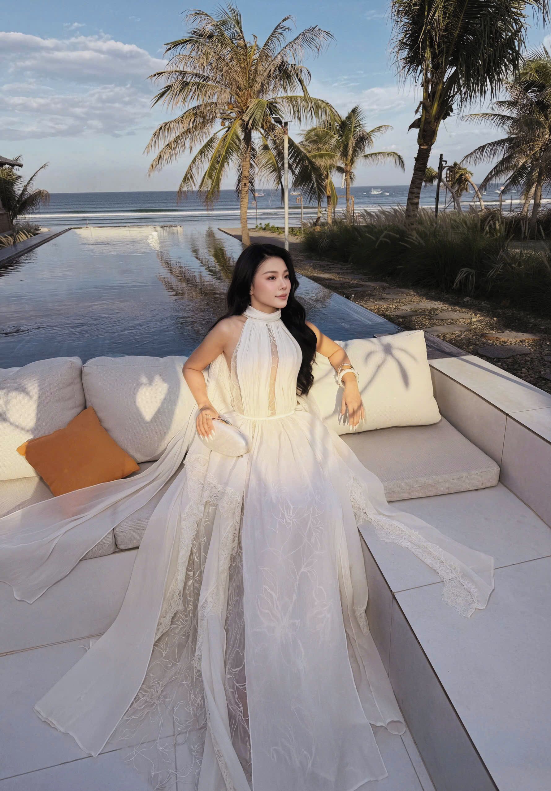 A person seated on a white outdoor sofa with a view of the ocean and palm trees.