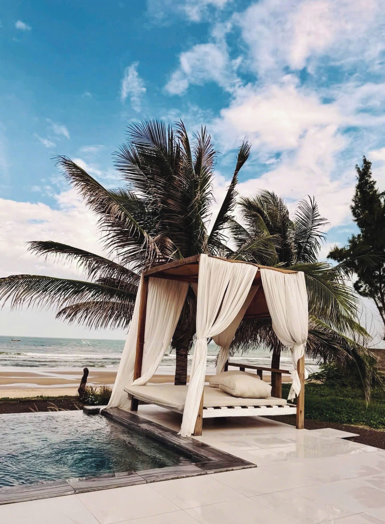 A beachside bed with a canopy, a pool, and palm trees.