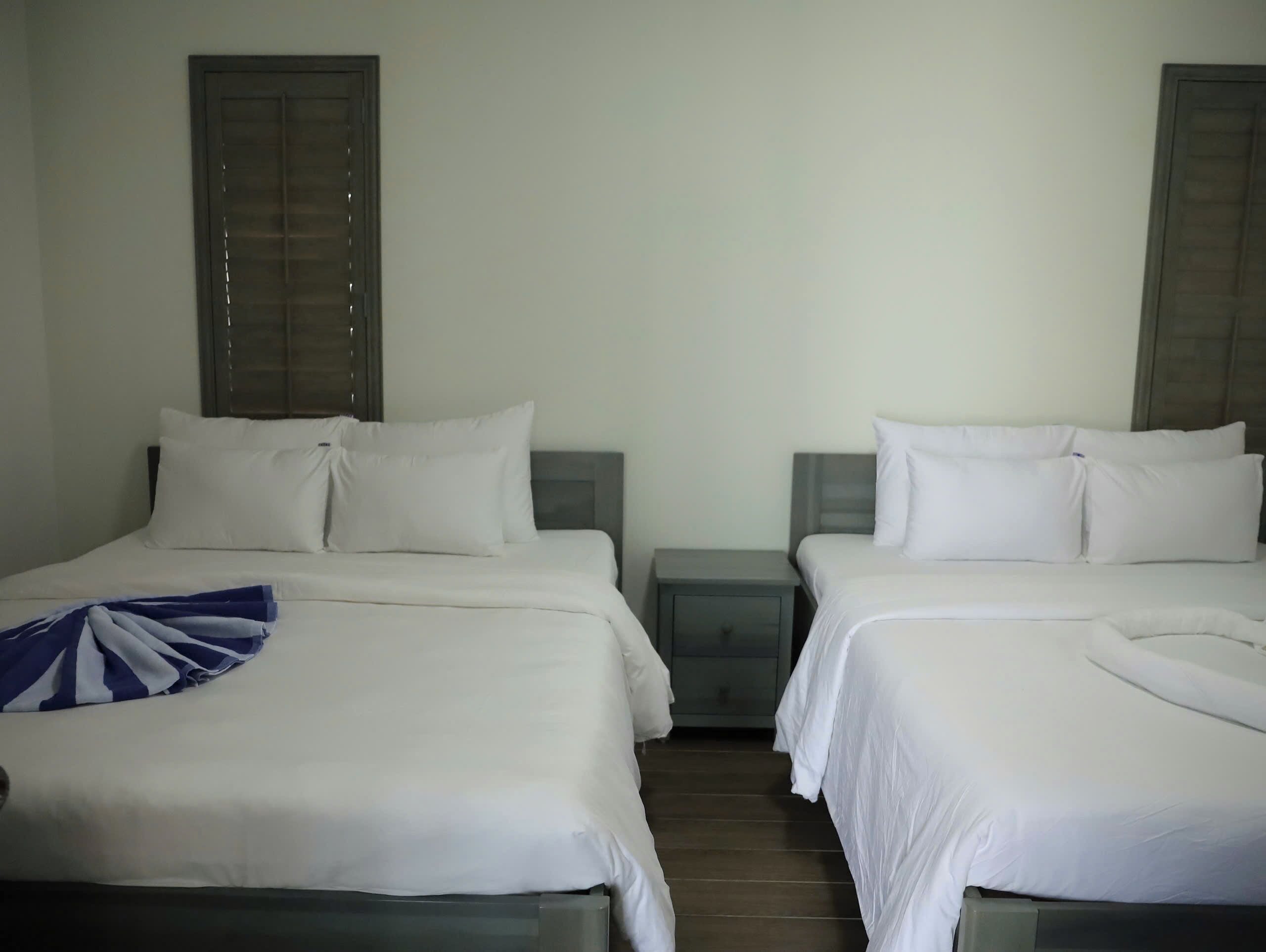 Quarto de hotel com duas camas, cada uma com roupa de cama e almofadas brancas, uma pequena mesa de cabeceira e uma janela com persianas.
