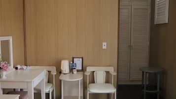 Una habitación con un panel de madera en la pared, un escritorio blanco con una silla, una mesita pequeña con una silla, un espejo y un armario cerrado con puertas persianas.