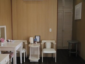 A room with a wooden paneled wall, a white desk with a chair, a small table with a chair, a mirror, and a closed closet with louvered doors.