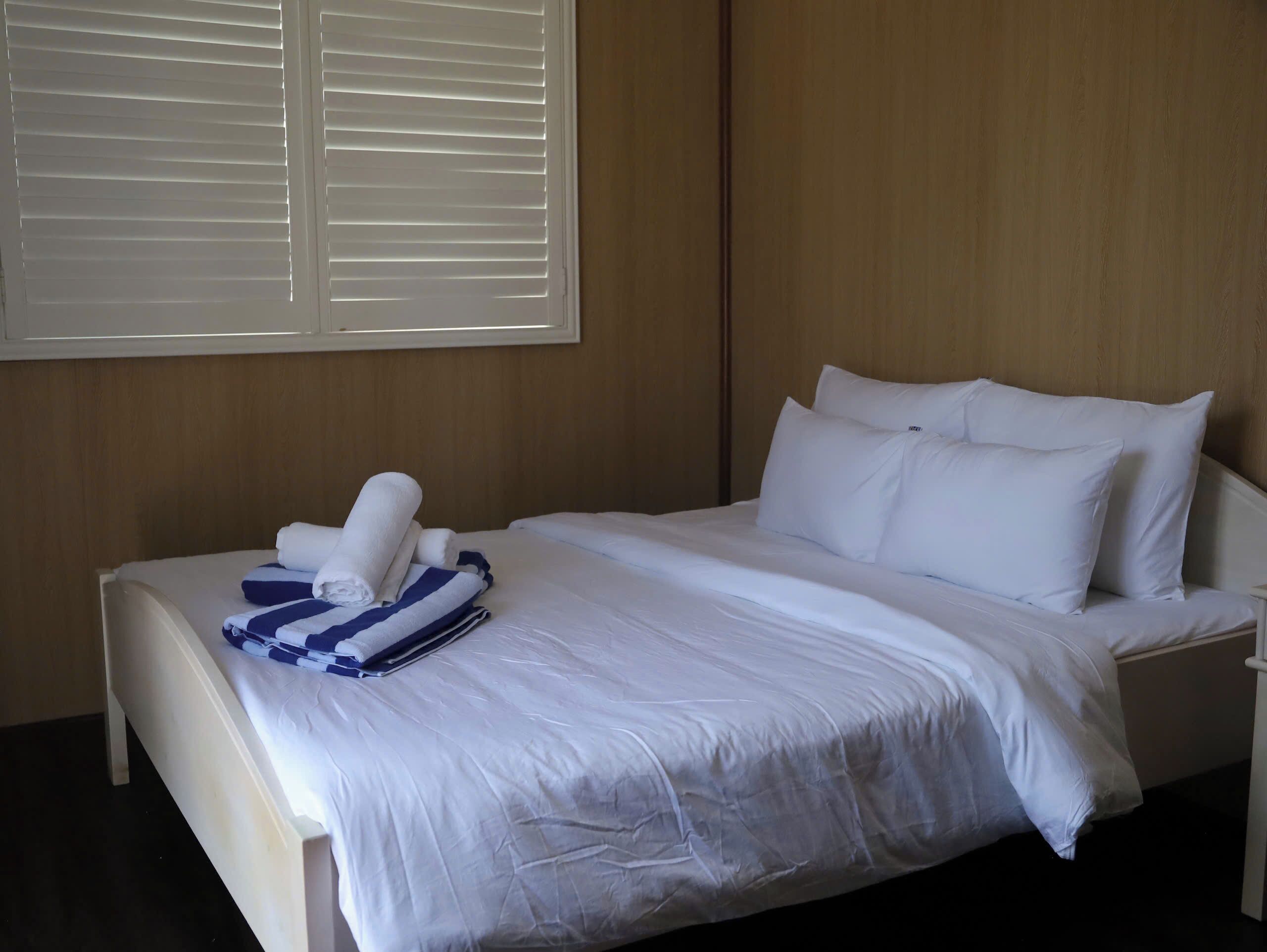 A neatly made bed with white linens and pillows, a blue and white striped towel, and a window with closed blinds.
