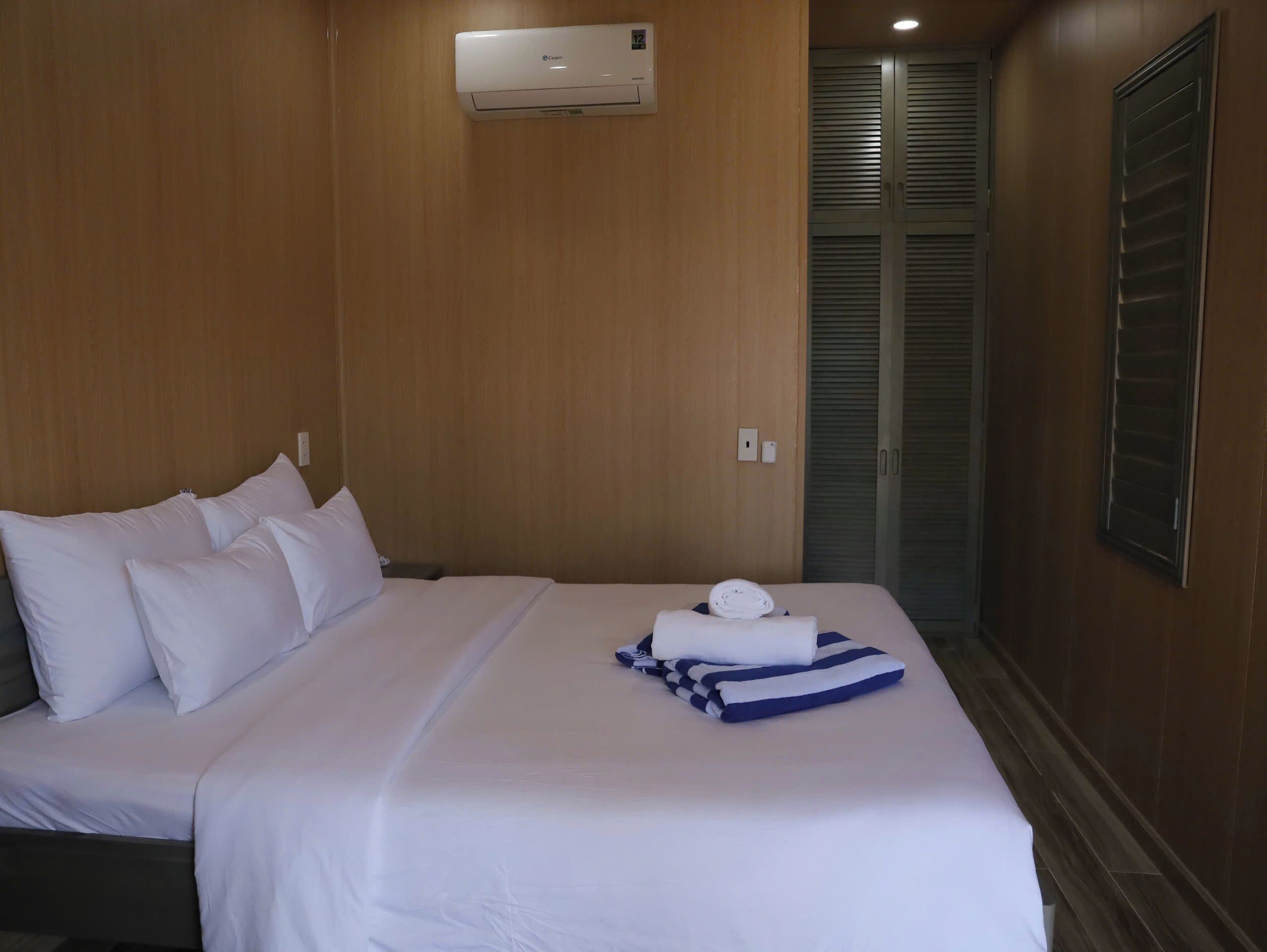 A hotel room with a bed, white linens, a folded towel, and a wall-mounted air conditioner.