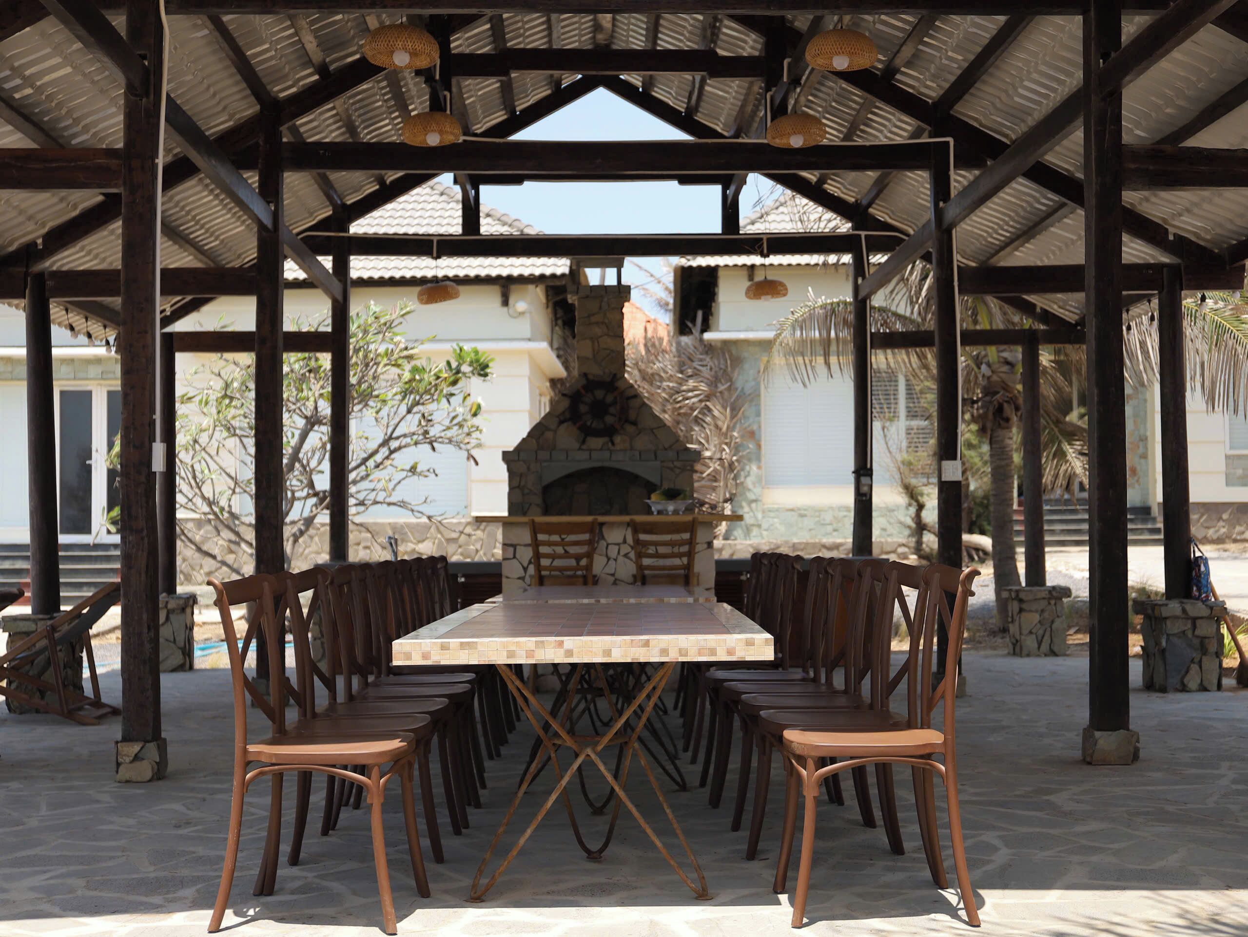 An outdoor covered dining area with a stone fireplace, wooden chairs, and a tiled table.