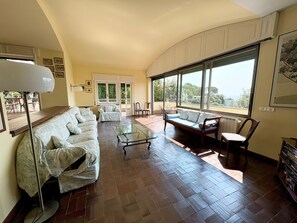 A spacious living room with a sofa set, a glass coffee table, and a view through large windows.