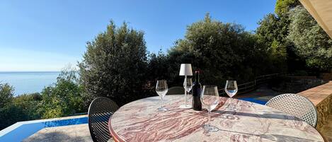 Una mesa redonda con cubierta de mármol, cuatro copas de vino, una botella de vino y una lámpara, frente a un cielo azul despejado y una masa de agua.