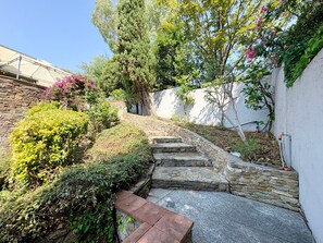 A stone staircase leading up to a white wall, surrounded by lush greenery and flowering plants.