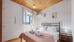 A bedroom with a wooden ceiling, a bed with a headboard, a bedside table with a lamp, and a window with a view. - Apartment 'Tolis House' (Kavallouri)