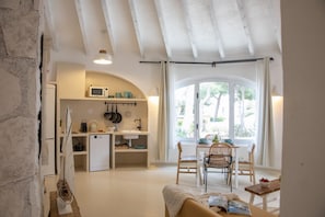 A compact kitchen with a microwave, sink, and various utensils. A dining area with a table and chairs is visible, and a window offers a view of greenery outside.