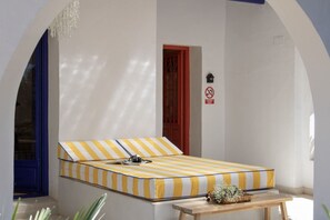 Ein Bett mit gelb-weiß gestreifter Bettwäsche, ein Nachttisch aus Holz mit einer Vase und eine rote Tür mit einem Rauchverbot-Hinweis.