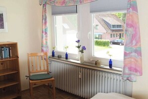 Ein Zimmer mit Fenster, Heizkörper, Holzstuhl und Bücherregal.